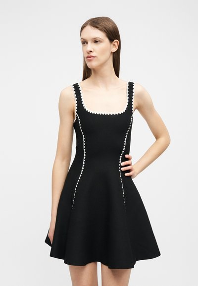 Femme portant une robe noire sans manches avec une bordure blanche en zigzag le long du décolleté et des côtés, debout avec une main sur la hanche.