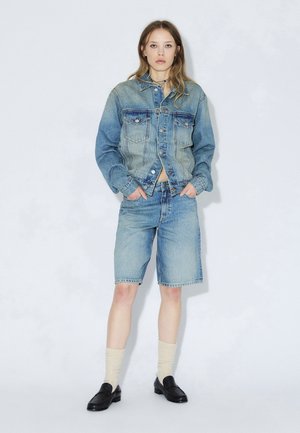 Lyseblå denimjakke med to brystlommer, kombineret med lyseblå denimshorts. Sorte loafers og cremefarvede knælange sokker fuldender looket.