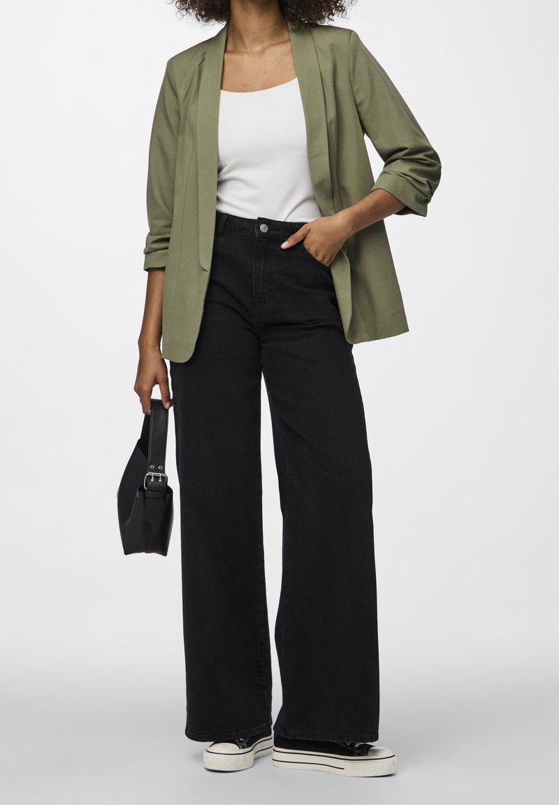 Femme portant un blazer vert olive, un haut blanc, un pantalon large taille haute noir, des baskets noires, tenant un petit sac à main noir.