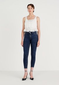 Witte, mouwloze top met een gerimpelde zoom en een trekkoord, gecombineerd met donkerblauwe skinny jeans en zwarte sandaaltjes met witte stippen.