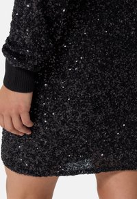 Abito nero in paillettes con superficie testurizzata, maniche aderenti e forma leggermente svasata all'orlo, che riflette la luce con accenti lucenti.