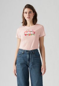 Rosa bomulls t-shirt med blommönster och texten "Levi's", matchad med blå vida denimjeans. Miljön är neutral och ljus.