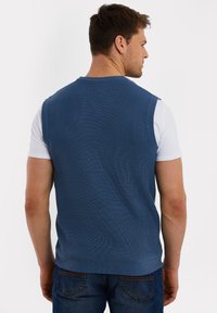 Blauwe geribbelde gebreide mouwloze trui, met ronde halslijn, gedragen over een wit T-shirt. Achteraanzicht toont naadloos ontwerp en gestructureerd materiaal.