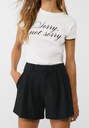 Kvinde iført en hvid t-shirt med teksten "Sorry, not sorry" og sorte højtaljede bukser med nålestriber, med den ene hånd i lommen.