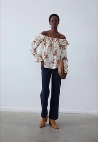 Blouse à épaules dénudées avec bords volants, fond crème et fleurs roses. Associée à un pantalon large bleu marine et des bottes à talons beige.