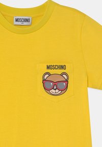 Žluté bavlněné tričko s grafikou medvěda s červenými slunečními brýlemi na kapsičce na hrudi. Název značky "Moschino" zobrazen nad kapsičkou.