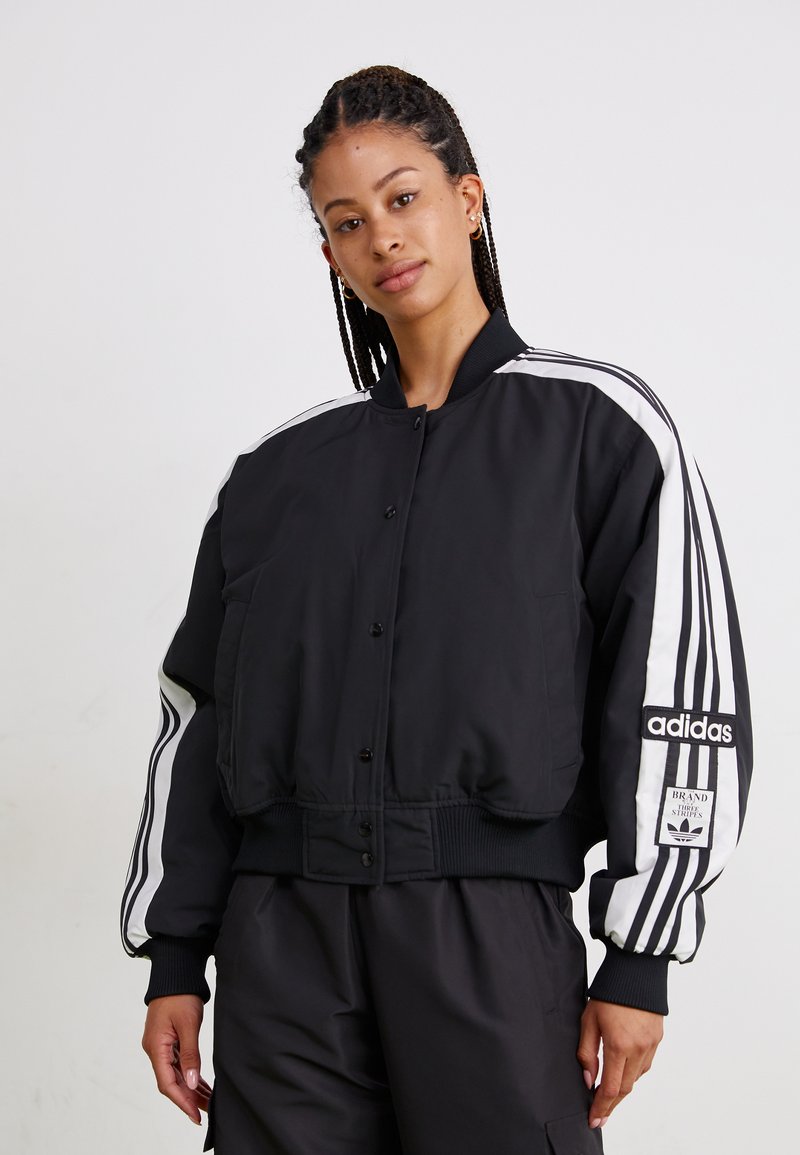Svart bomberjakke med hvite striper nedover ermet, trykknapper og ribbede mansjetter. Har et Adidas-logo-patch på ermet.