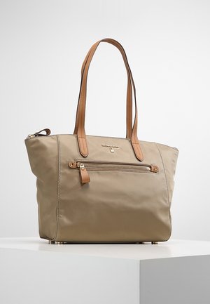 Béžová taška přes rameno s hnědými koženými madly, přední kapsou na zip a zlatým logem značky na bílém povrchu proti světlému šedému pozadí.