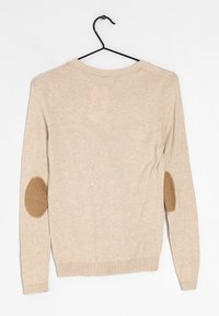 Beige Strickpullover mit langen Ärmeln, geripptem Saum und Ellenbogen-Patches aus strukturiertem Stoff. Hängt an einem schwarzen Haken vor einem weißen Hintergrund.