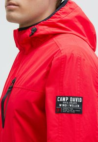 Veste à capuche rouge en matériau léger. Dotée d'une étiquette noire sur la manche droite avec un logo et du texte. Poche zippée à l'avant.