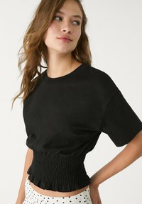 T-shirt in cotone nero con scollatura rotonda e maniche corte. Presenta una vita voluminosa con orlo arricciato. Pantaloni corti a pois parzialmente visibili.
