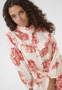 Light fabric blouse featuring a cream background with large red floral patterns, ruffle accents, and button closure. Long, puffed sleeves.