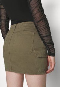 Olivengrønn denim miniskjørt med to baklommer og én side cargo-lomme. Har en ren finish og solid søm.