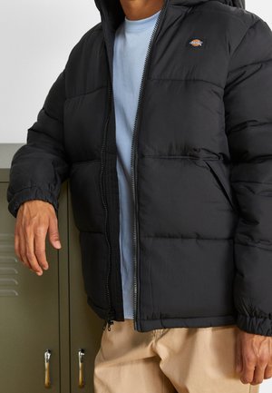 Puffer jacket in black with a zip front, hood, and elastic cuffs. Features a small logo on the chest and a textured fabric.