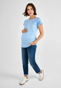 Ljusblå kortärmad gravidtröja kombinerad med blå jeans och vita sneakers. Tröjan har en avspänd passform och rund halsringning.