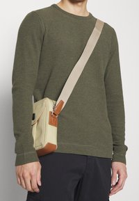 Sac bandoulière beige avec des accents en cuir marron, fermeture éclair et sangle réglable, porté sur une chemise à manches longues texturée vert olive.