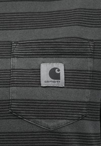 šedá pruhovaná tkanina s kapsou, na které je čtvercová šedá značka. Značka zobrazuje logo "Carhartt" v černé a bílé.