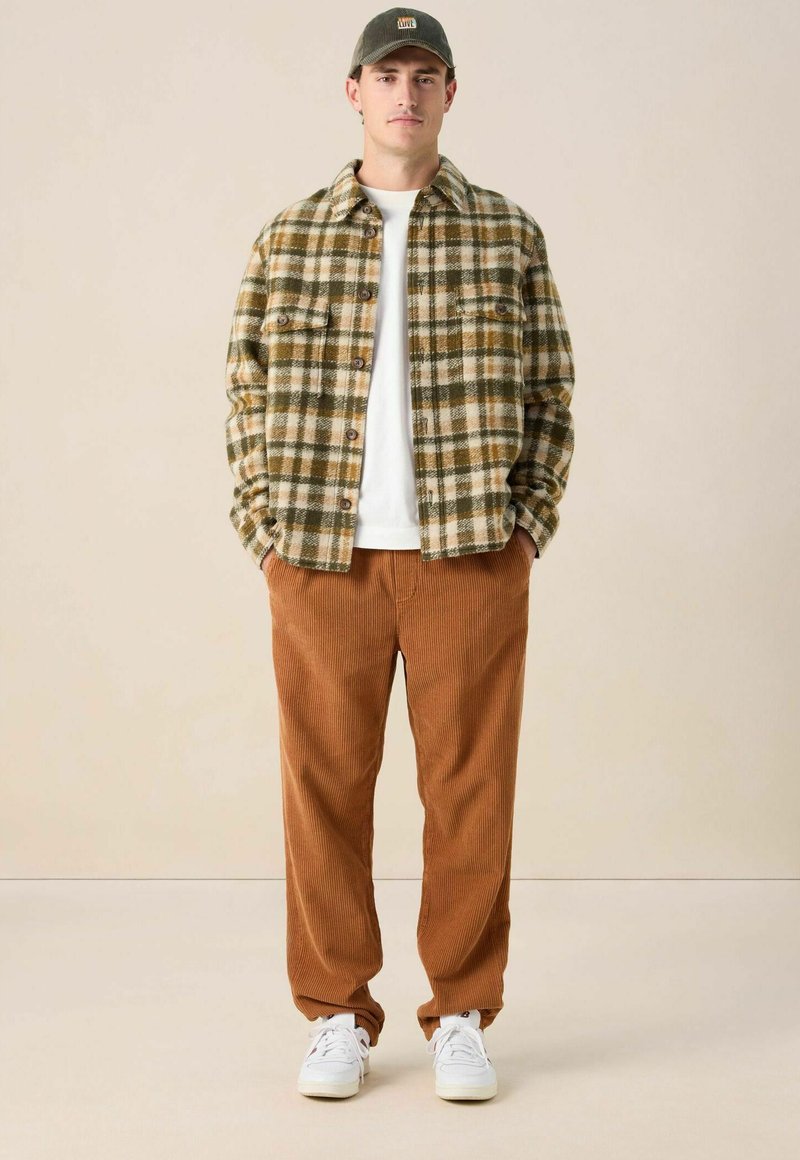 Homme portant une veste à carreaux verte et marron, un t-shirt blanc, un pantalon en velours côtelé marron, des baskets blanches et une casquette vert olive, se tenant les mains dans les poches.