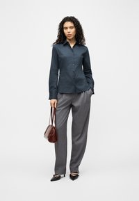 Γυναίκα με κατσαρά μαλλιά που φοράει σκούρο πουκάμισο με κουμπιά, γκρι παντελόνι, μαύρες γόβες, κρατώντας καφέ τσάντα, στέκεται μπροστά σε απλό λευκό φόντο.