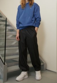 Osoba s dlouhými kudrnatými vlasy, oblečená v modré mikině, černých širších kalhotách a bílých teniskách, stojí blízko vnitřního schodiště se skleněným zábradlím.