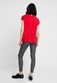 Mujer de pie, de espaldas, vestida con una blusa roja de manga corta, pantalones ajustados de rayas verticales en blanco y negro, y zapatos negros con cordones.