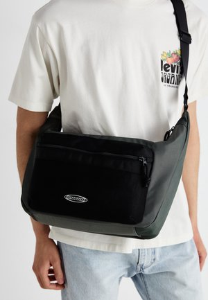Shoulder bag in black and dark green materials, featuring a textured front pocket, zipper closure, and logo detail. Smooth, casual design.