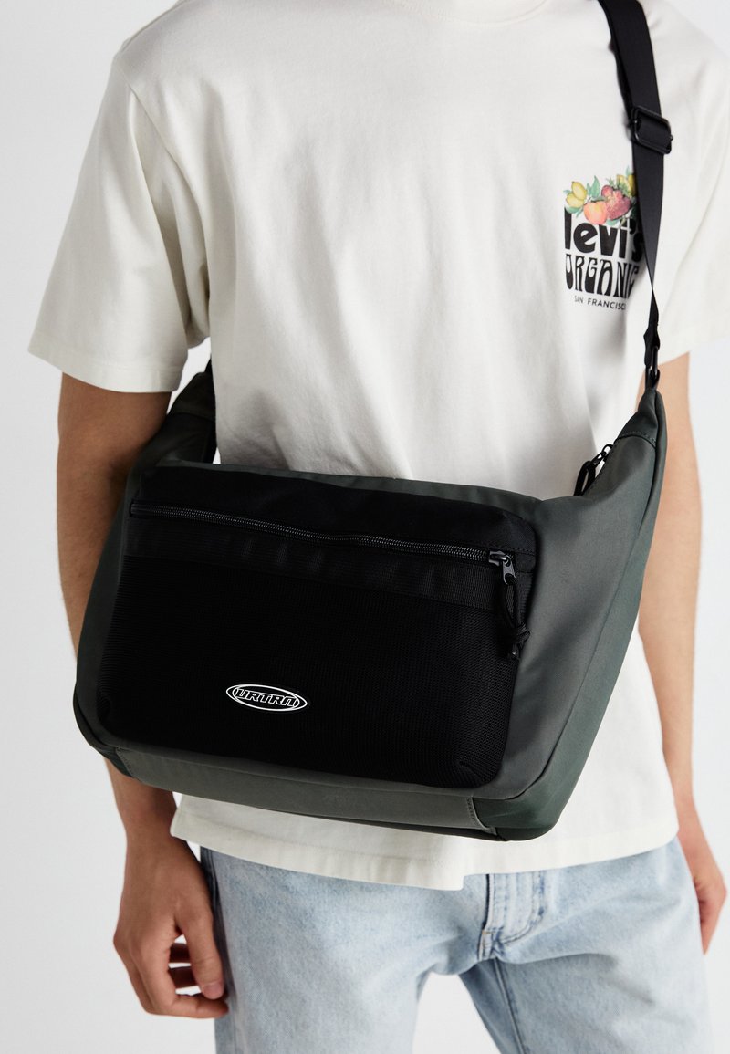 Bolso de hombro en materiales negro y verde oscuro, con un bolsillo delantero texturizado, cierre de cremallera y detalle de logo. Diseño liso y casual.