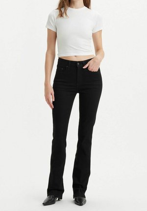 Vrouw draagt een witte korte top met korte mouwen, zwarte hoge taille jeans en zwarte laarzen met spitse neus, staand tegen een effen achtergrond.