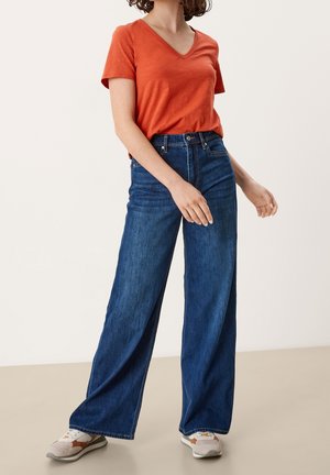 T-shirt orange à col en V associé à un jean bleu taille haute et coupe large. Les jeans présentent une finition denim classique, des coutures discrètes et des poches.