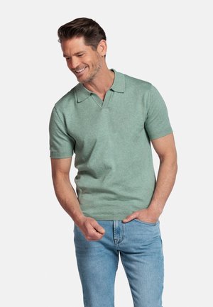 Man draagt een lichtgroen poloshirt met korte mouwen en lichtblauwe spijkerbroek, glimlacht en kijkt naar beneden met één hand in de zak.
