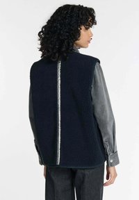 Marineblauer Fleece-Weste mit strukturiertem Oberflächen, ärmellosem Design und einem kontrastierenden silbernen Streifen entlang der Mitte des Rückens.