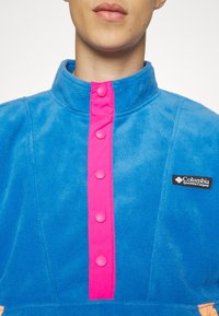 Pullover in pile di un vivace blu con colletto alto, dotato di una chiusura con bottoni rosa brillante e etichetta con logo Columbia sul petto.