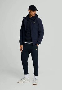 Jachetă de tip zip-up de marină cu glugă peste un hanorac negru, asociată cu pantaloni de trening de marină și adidași gri, având un mic accent cu logo galben.