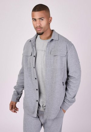 Chemise grise à boutons, fabriquée en tissu doux, dotée de deux poches poitrine et d'un col, portée par-dessus un t-shirt gris assorti.