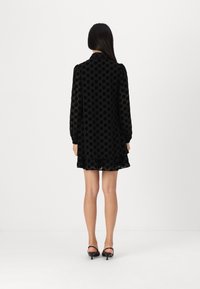 Robe noire à pois avec de longues manches bouffantes, en matériau transparent, et une jupe évasée. La vue arrière montre de légers volants au niveau de la jupe.