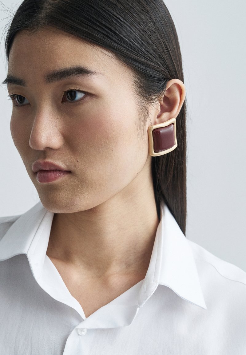 Vierkante bordeauxoorbel met een gouden omlijsting, met een glad oppervlak. Het model draagt een witte blouse met kraag.