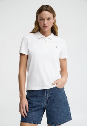 Kvinde iført en hvid poloshirt med korte ærmer og en lille sort logo samt blå denimshorts, stående med den ene hånd i lommen.