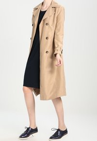 Béžový trench coat s dvojitým zapínáním, velkými klopy a knoflíkovými detaily, nošený přes černé šaty a doplněný tmavými botami.