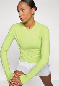 Długi rękaw, jasna limonkowa zielona sportowa bluza z lekkiego materiału; posiada okrągły dekolt i mankiety z otworami na kciuki. Biały logo Nike na klatce piersiowej.