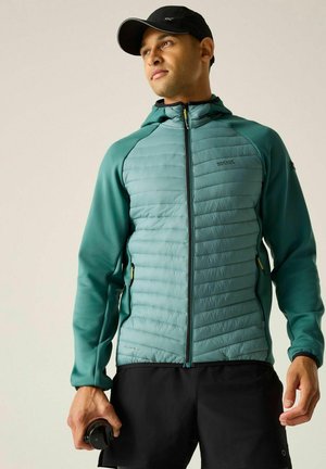 Uomo con giacca trapuntata turchese, pantaloncini neri e cappellino nero, che tiene un oggetto nero nella mano destra, guardando di lato su sfondo uniforme.