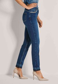 Dunne blauwe jeans met een medium hoge taille, met achterzakken en een kettingaccent, gecombineerd met beige hoge hakken.