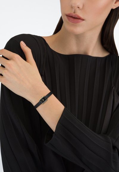 Fekete hatszögletű óra arany színű részletekkel, sötét szíjon. Egy kézen viselik, amely egy redős fekete ruhadarab ellen látható. Minimalista kialakítás.