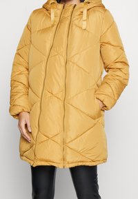 Gul kviltad pufferjacka med huva, utrustad med diagonala sömmar, dragkedjestängning och sidofickor. Slätt och lätt tyg.