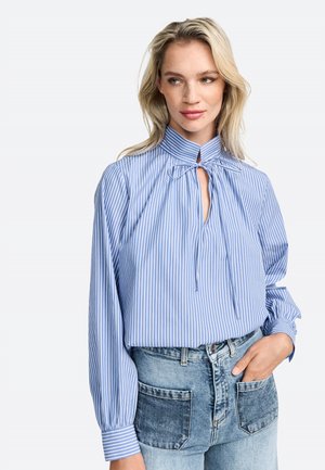Blouse rayée bleu et blanc avec un col haut, un détail à nouer au cou et de longues manches bouffantes, associée à un jean en denim taille haute.