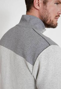 Grau und hellgrauer Pullover mit hohem Kragen, der ein strukturiertes Strickmuster, ein Farbabstufungsdesign und sichtbare Nähdetails an der Schulter aufweist.