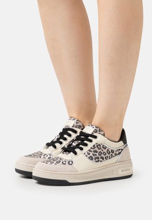 Jambes portant des baskets basses beige et noires avec des panneaux en imprimé léopard, des lacets noirs et le logo "GUESS" sur le côté de la semelle.