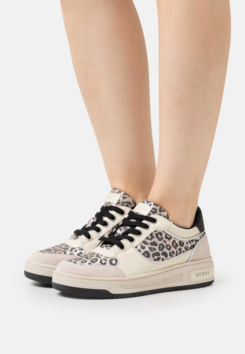 Jambes portant des baskets basses beige et noires avec des panneaux en imprimé léopard, des lacets noirs et le logo "GUESS" sur le côté de la semelle.