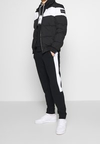 Muž v černé a bílé prošívané bundě, černé mikině s kapucí, černých teplákových kalhotách s bílými bočními pruhy a bílých teniskách, stojí proti jednobarevnému pozadí.