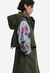 Veste vert olive au design ajusté avec capuche, présentant une manche grise décorée d'un graphique coloré de Minnie Mouse et d'accents floraux.