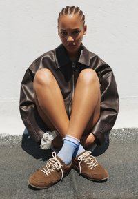 Giacca in pelle marrone con vestibilità oversize, abbinata a sneakers in camoscio marrone e calze azzurre chiare. Modello seduto con le gambe incrociate sul pavimento.